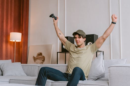 A young man joyfully celebrates winning a video game, sitting on a couch with his arms raised in triumph.